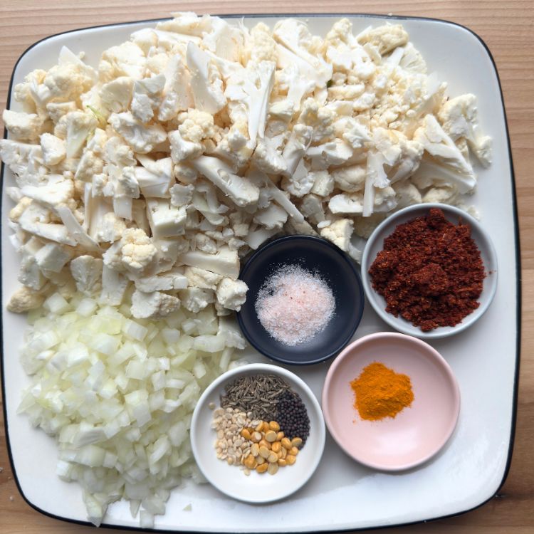 Top view of all ingredients for Cauliflower Vepudu Fry, including cauliflower, onion, spices, and tempering seeds, arranged on a plate.