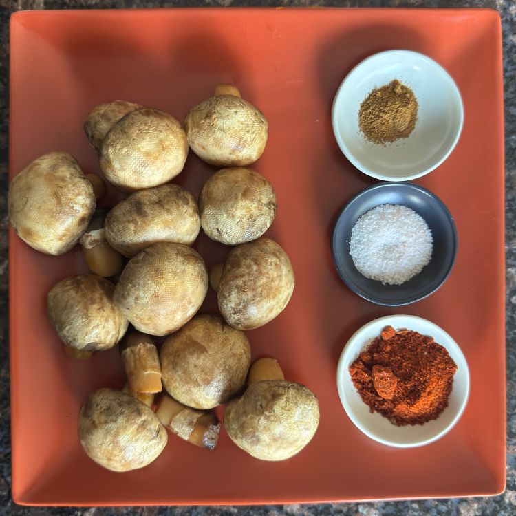 Fresh ingredients for Indian spicy mushroom fry including button mushrooms, chili garlic powder, and cumin powder arranged on a rustic plate