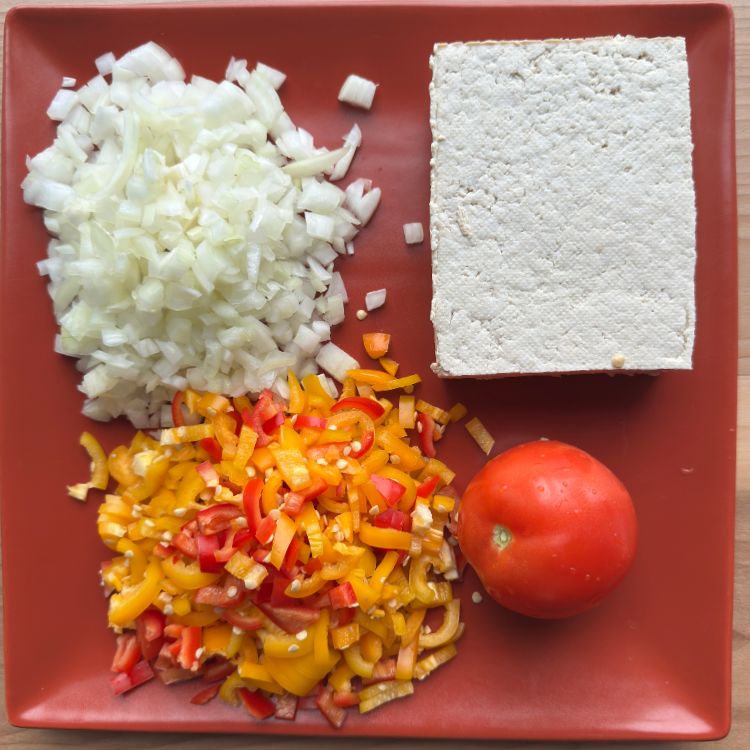 Fresh ingredients for Tofu Burji including crumbled tofu, onions, tomatoes, and peppers laid out on a rustic wooden surface